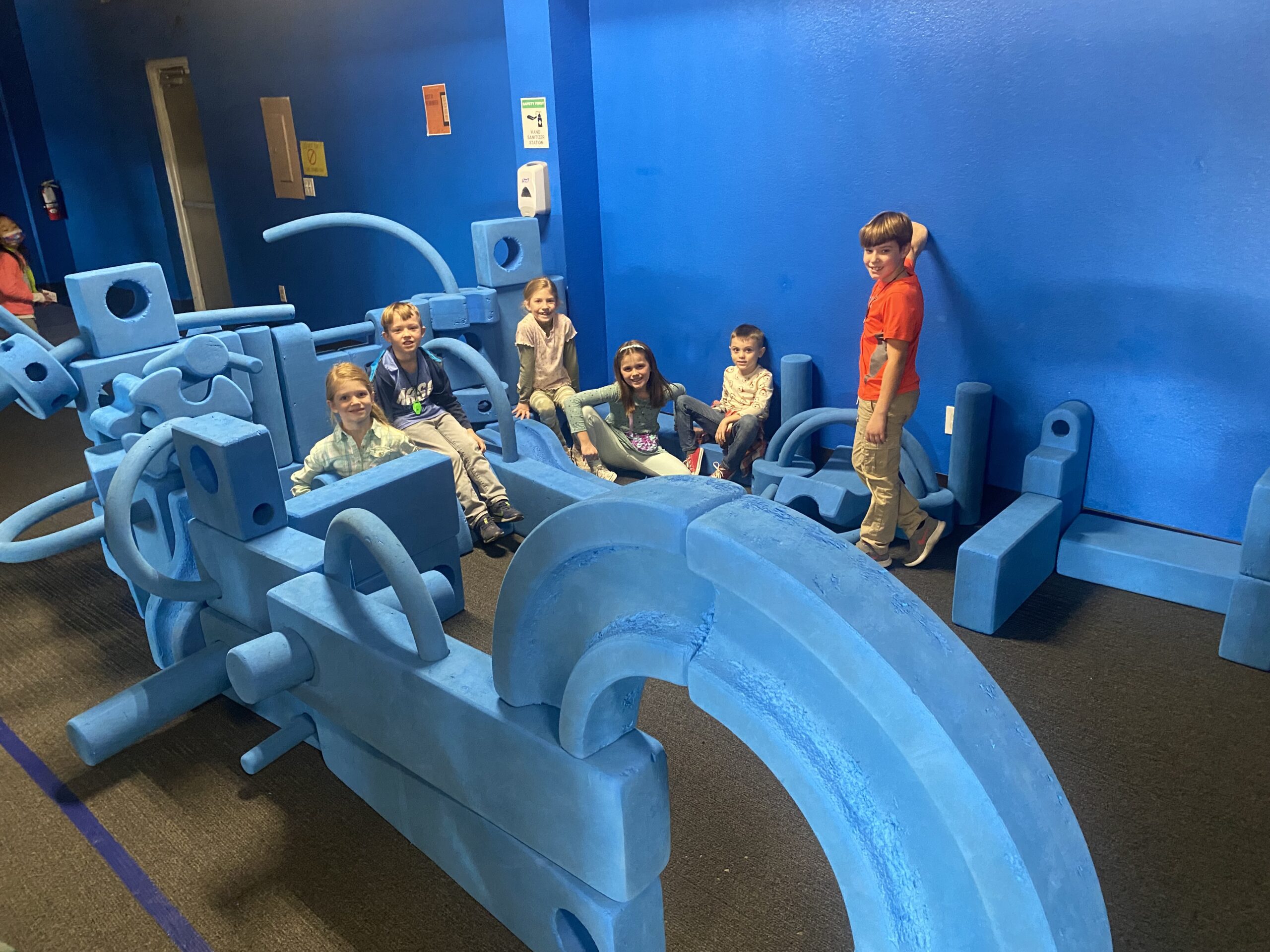 Several children playing on and around a large, elaborate structure built from blue foam playground blocks inside a children's museum.