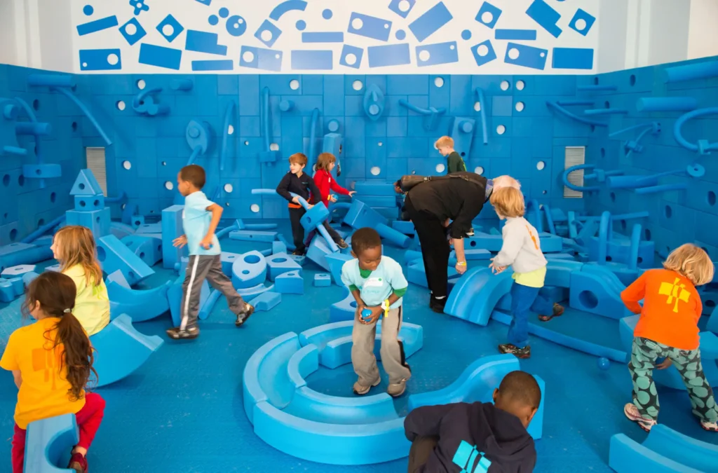 A vibrant indoor play space featuring a Custom Foam Block Wall and Big Blue Blocks. Children are seen interacting with the modular foam wall and large building blocks, showcasing how the interactive play space encourages architectural-scale construction and collaborative play.