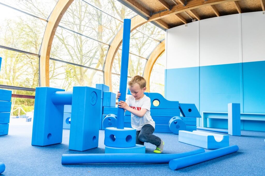 Children collaborating and building a custom structure using large blue foam Imagination Playground blocks in an outdoor setting.