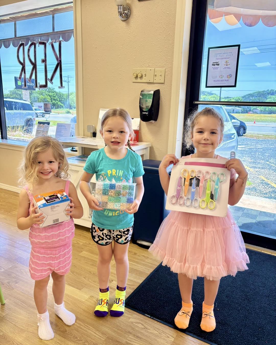 Three young girls happily posing together in a brightly lit playroom, showcasing their craft supplies.