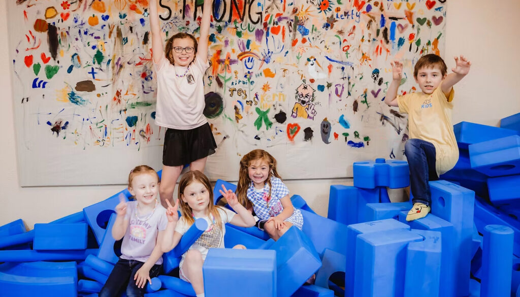 A group of young children smiling and playing together with large blue foam building blocks in an indoor classroom setting at Kerr Kids.
