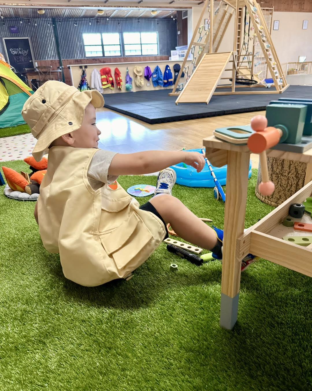 A young child happily playing on a wooden and rope swing structure  on an artificial grass lawn, enjoying an active open-ended play session.