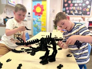 Preschool children collaborating at a classroom table to assemble an Imagination Playground Dino Bones dinosaur skeleton next to a tall blue block tower structure.