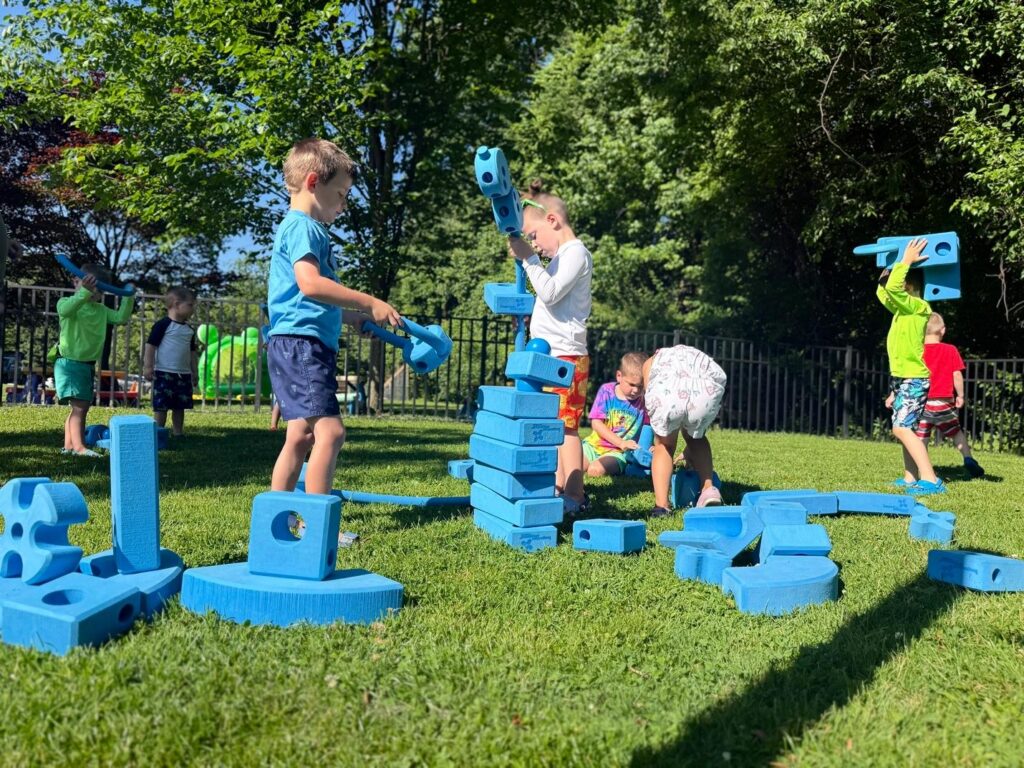 Several young children playing together outdoors on a sunny day, using large blue foam building blocks to construct shapes and steps on a grassy lawn.