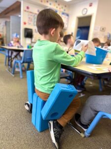 A young preschool boy sitting at a classroom desk, focusing intently as he constructs a custom design with blue foam building blocks.