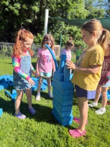 Three young preschool children playing together outdoors on a sunny day, collaborating to build a tall tower using Imagination Playground large blue foam blocks.
