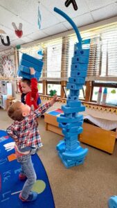 A young preschool boy reaching up high to carefully balance a piece on top of a tall vertical tower structure made from blue foam blocks in a classroom.