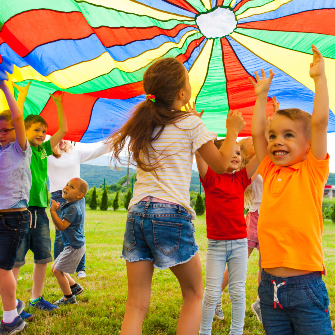 A diverse group of young children laughing and cheering while holding up a vibrant, multicolored parachute on a sunny, grassy field.